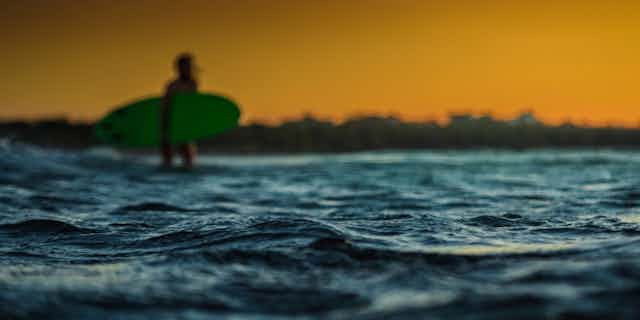 A blurred photo of a surfer with a green surfboard. The image focuses on the rippling sea in the foreground. 