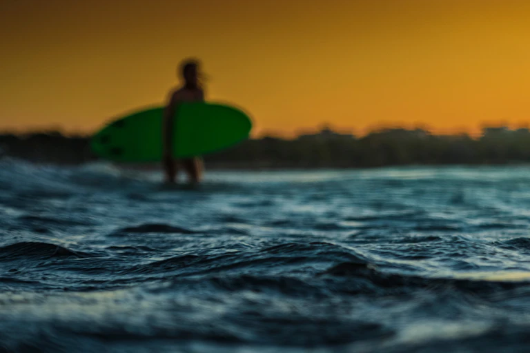 A blurred photo of a surfer with a green surfboard. The image focuses on the rippling sea in the foreground.