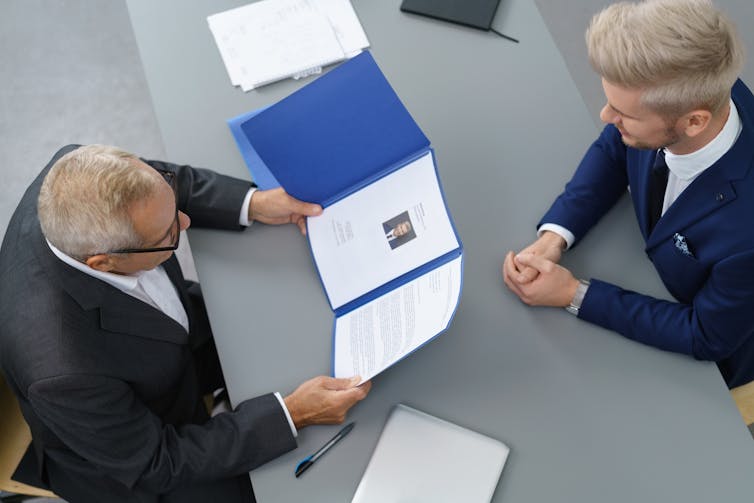 Overhead shot of two men in suits facing each other across a desk, one holding an open file with documents.