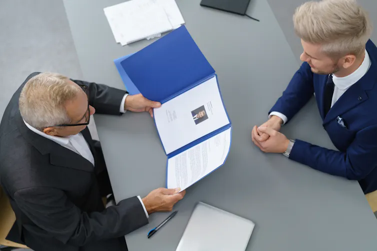 Overhead shot of two men in suits facing each other across a desk, one holding an open file with documents.