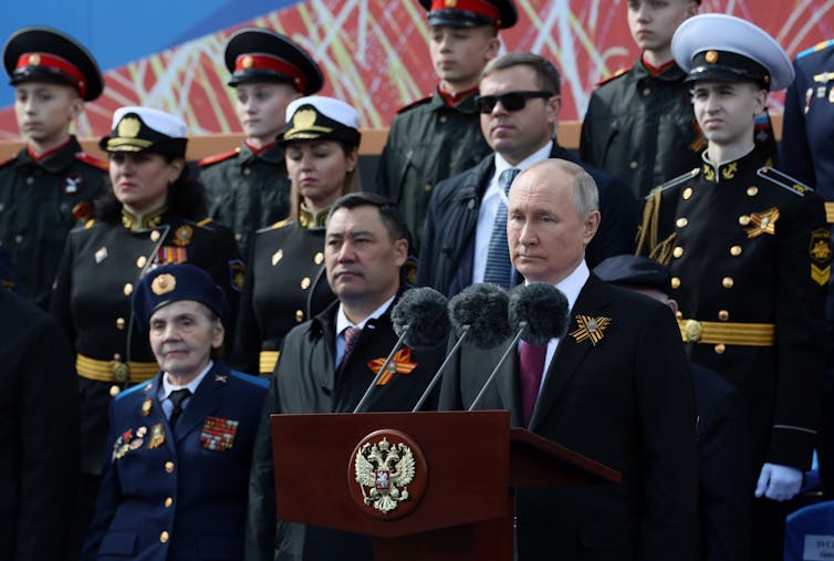 Vladimir Putin stands behind a row of microphones surrounded by aides and military officers.
