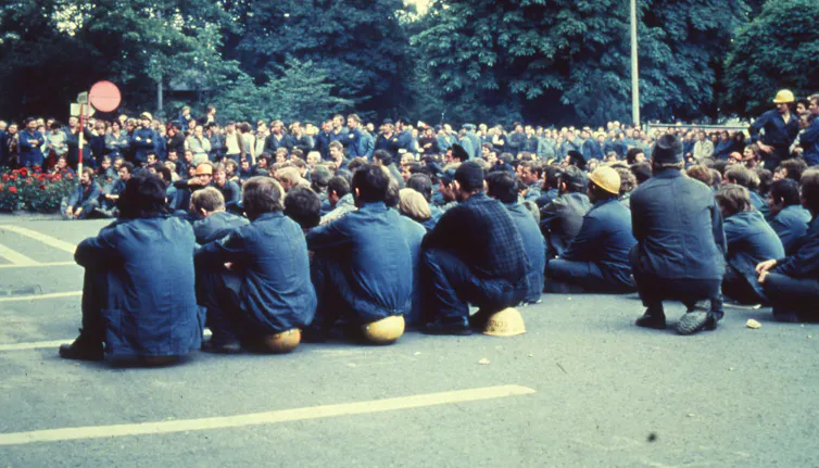 Men in blue sit down on hard hats on the ground.