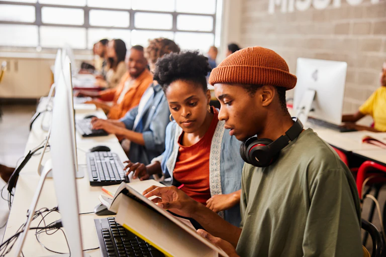 Young people work in a computer lab, chatting and sharing textbooks as they do so
