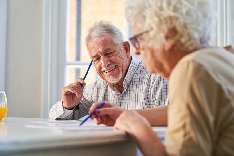 An elderly man holding a pencil smiles while completing a written task. An elderly woman sitting beside him writes on a piece of paper.