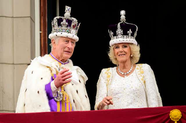 An elderly man and women wear large ornate crowns and robes as they stand on a balcony with red bunting.