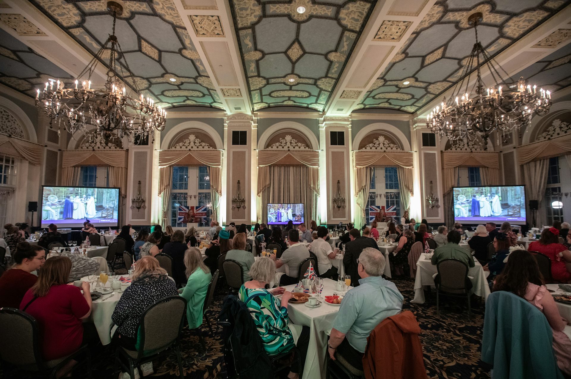 People sit in an ornate ballroom drinking tea with two TV screens showing the coronation at the front of the room.