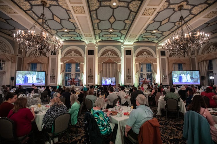 People sit in an ornate ballroom drinking tea with two TV screens showing the coronation at the front of the room.