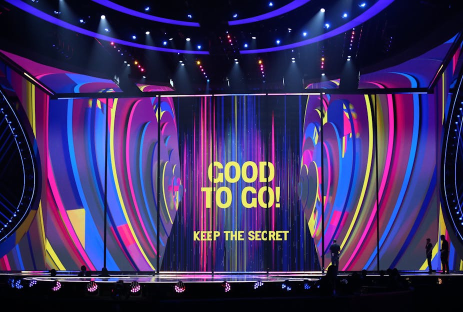 The words GOOD TO GO! KEEP THE SECRET appear on a stage in yellow. The background is swirls of blue, pink and purple. 