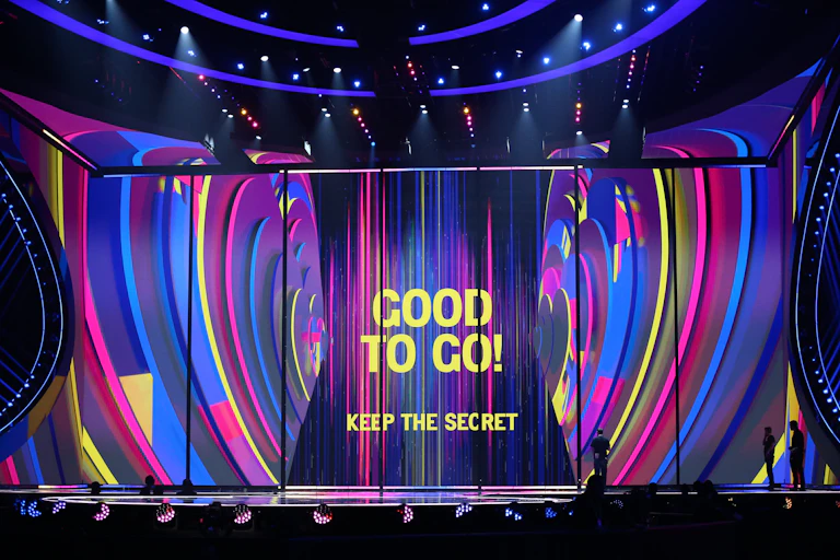 The words GOOD TO GO! KEEP THE SECRET appear on a stage in yellow. The background is swirls of blue, pink and purple.