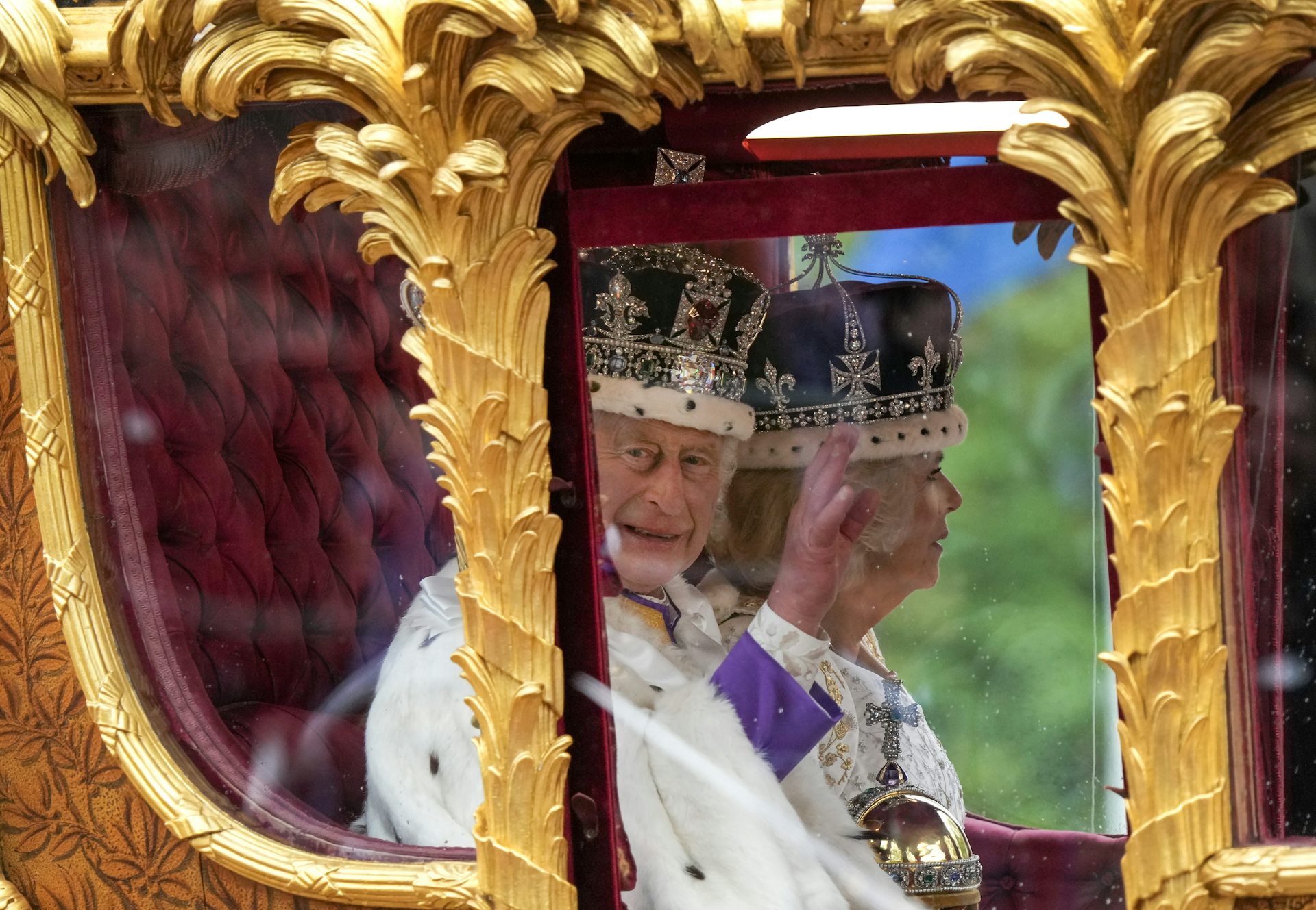 An elderly man in a large crown adorned with jewels and purple velvet waves from an ornate golden horse-drawn carriage. An elderly woman in a similar crown sits beside him.