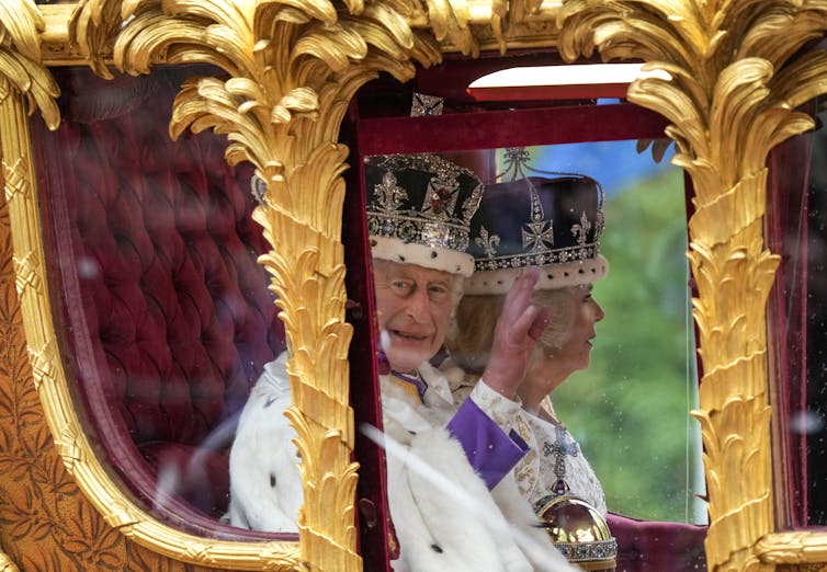 An elderly man in a large crown adorned with jewels and purple velvet waves from an ornate golden horse-drawn carriage. An elderly woman in a similar crown sits beside him.