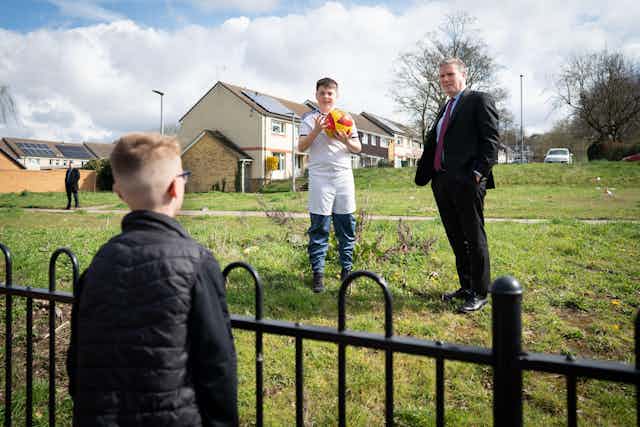 A boy talking over a fence to Keir Starmer, who is standing next to another boy holding a ball.