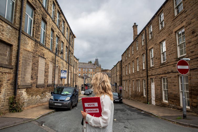 A woman holding a clipboard reading 'vote Labour' looks down a street of boarded up buildings.