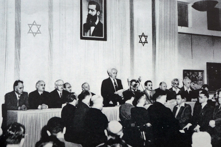 A white haired man in a dark suit and tie stands reading from paper in his hand, flanked by nearly a dozen people.