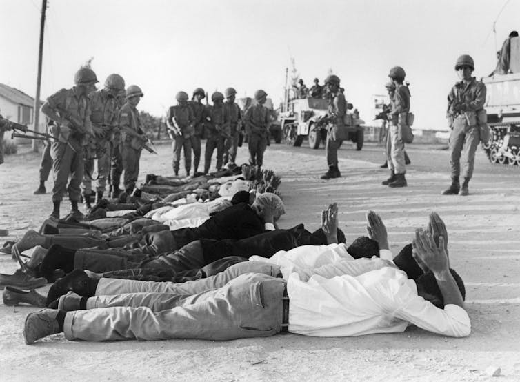 Men lying down on the ground with their hands behind their heads, overseen by armed soldiers.