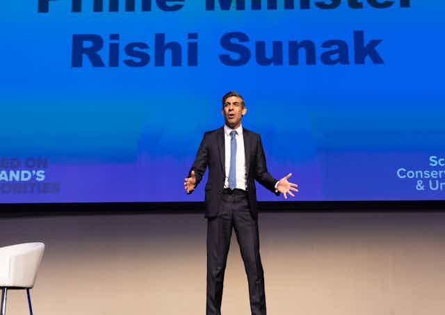 Rishi Sunak giving a speech in front of a huge screen bearing his name.