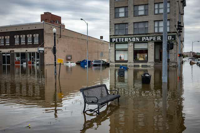 A flooded business district with a paper company  building flooded up to its windows and an empty metal bench outside. Several cars are underwater in the background.