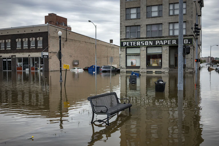 A flooded business district with a paper company building flooded up to its windows and an empty metal bench outside. Several cars are underwater in the background.