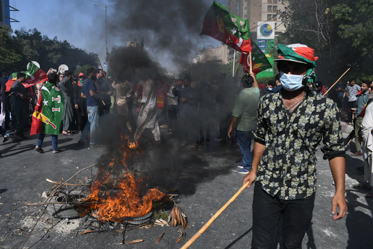 A man in a mask holding a long stick stands by a fire.