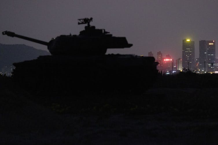 A night scene of a silhouetted tank with lit-up skyscrapers in the distance.