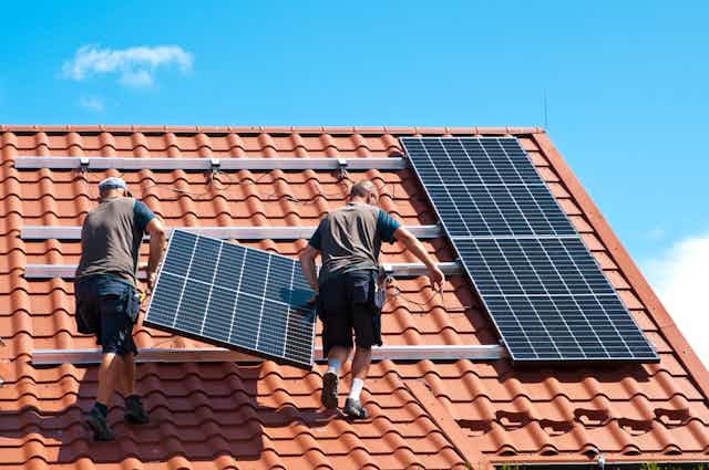 Two installers carry solar panels up a tiled roof