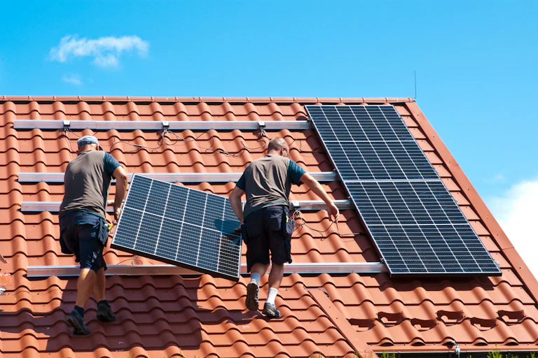 Two installers carry solar panels up a tiled roof
