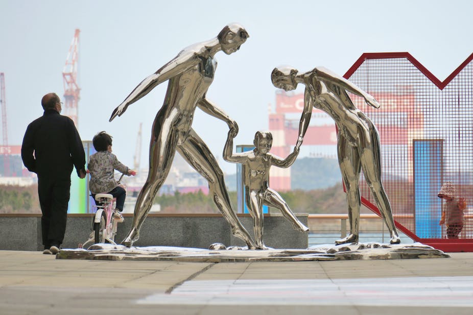 An elderly man guides a child on a bicycle nex to statues depicting a family