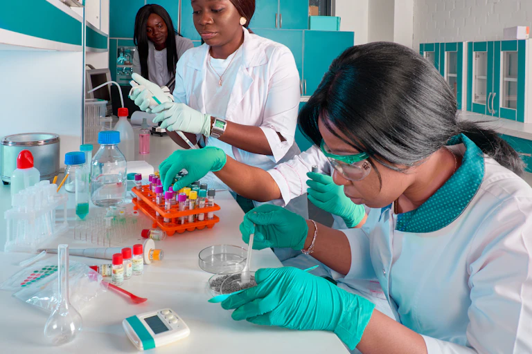 Three women in laboratory coats, goggles and gloves work with vials and Petri dishes