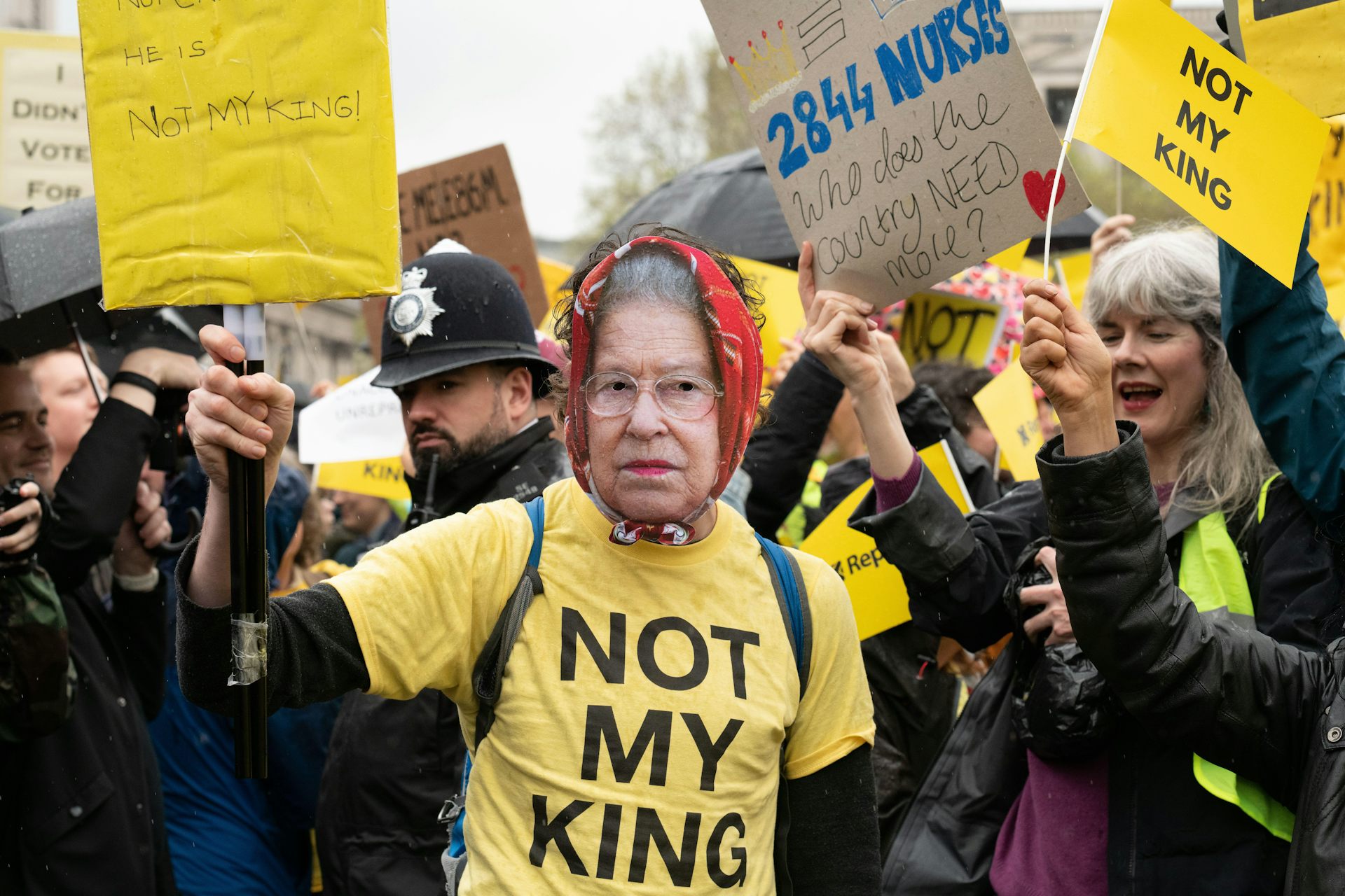 A protester wearing a mask of queen elizabeth II and a yellow t shirt with large black text reading 'not my king'