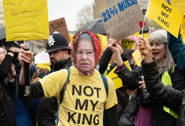 A protester wearing a mask of queen elizabeth II and a yellow t shirt with large black text reading 'not my king'