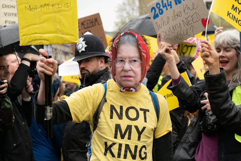 A protester wearing a mask of queen elizabeth II and a yellow t shirt with large black text reading 'not my king'