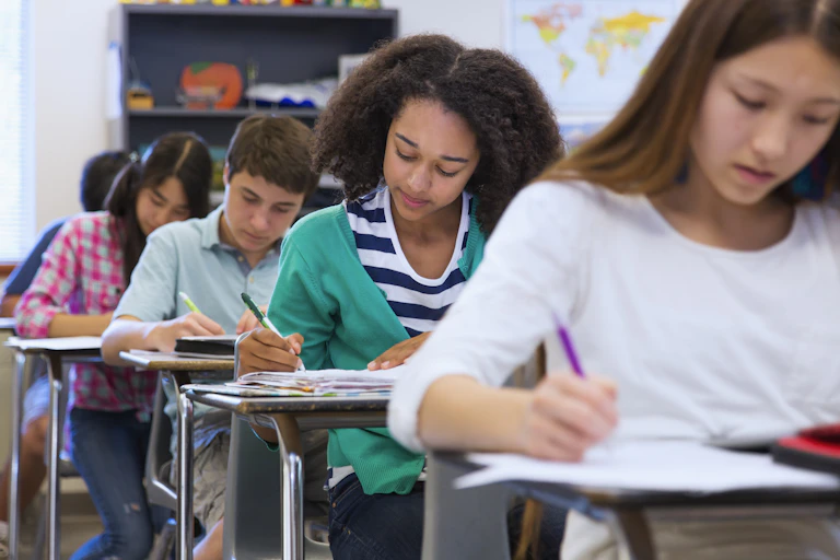 A column of diverse students sit at their desks working on assignments as a world map hangs in the background.