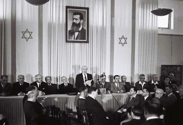 David Ben-Gurion declaring independence beneath a large portrait of Theodor Herzl, founder of modern Zionism