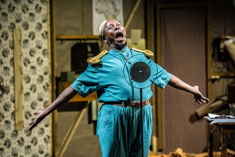 A man in a bright turquoise military outfit sings, his arms outstretched, into a vintage microphone.