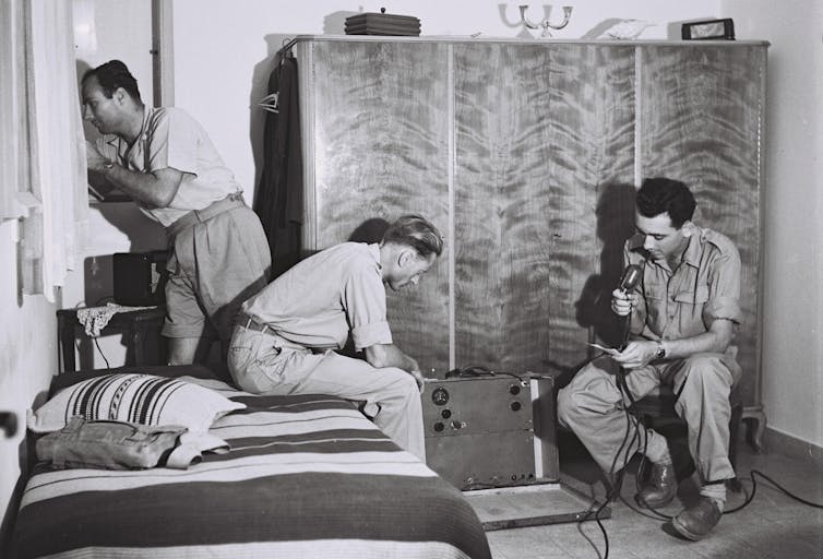 Black and white photo of three men in a small bedroom, one speaking into the microphone of a large radio device.