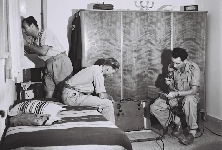 Black and white photo of three men in a small bedroom, one speaking into the microphone of a large radio device.