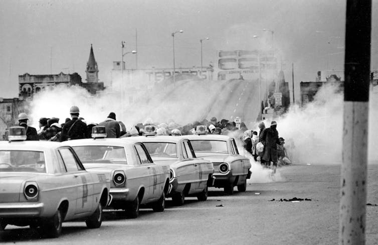 A black-and-white photo sows a line of police cars and clouds of tear gas.