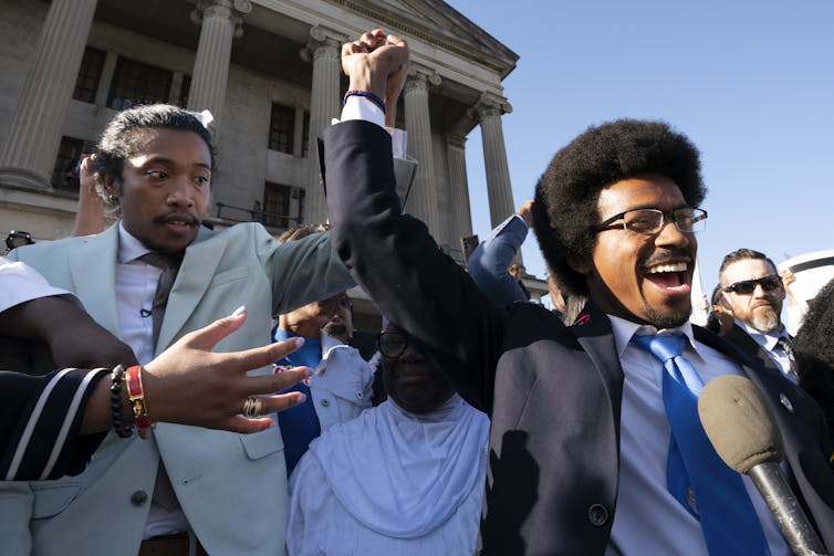 Two black men join hands and raise them over their heads surrounded by supporters in front of a legislative building.