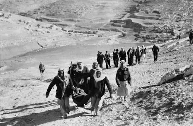 Two men in Arab dress carrying what appears to be a wounded person lead a line of men and women walking through a desert.