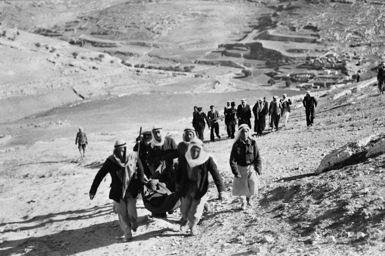 Two men in Arab dress carrying what appears to be a wounded person lead a line of men and women walking through a desert.