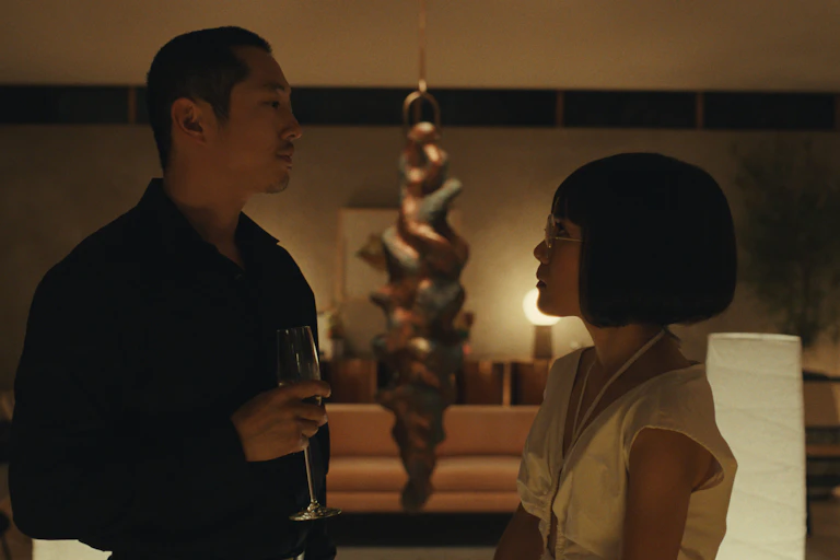 Danny and Amy sip champagne, a large art sculpture hangs behind them.