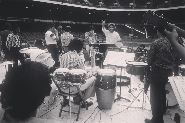 Un hombre delante de una orquesta latina hablando con el brazo en alto.