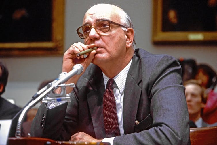 Paul Volcker, glasses, suit & tie, cigar; in front of microphone, seated at a desk, people and paintings in the background.