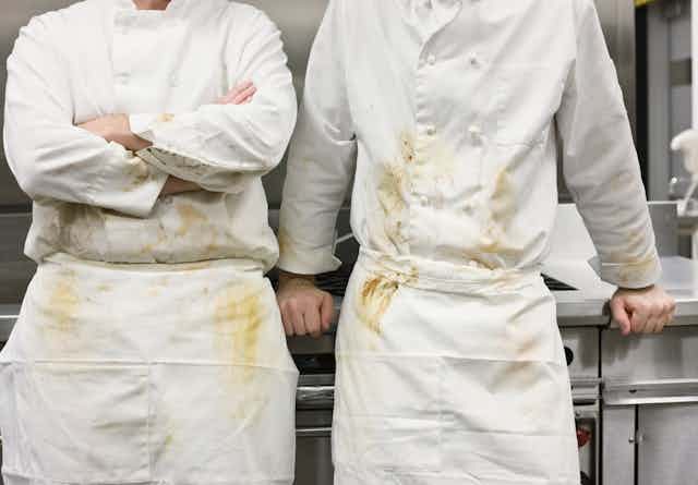 Two chefs with food-stained smocks lean against a stove.