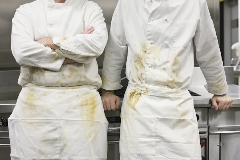 Two chefs with food-stained smocks lean against a stove.