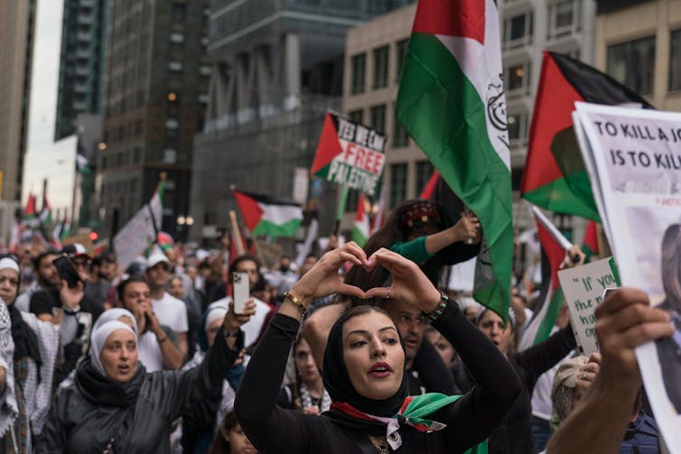 A woman making a heart shape with her hands fronts a throng of people waving Palestinian flags.