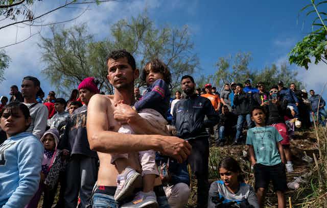A barechested man holds his daughter as they join dozens of others waiting to receive asylum in the U.S.