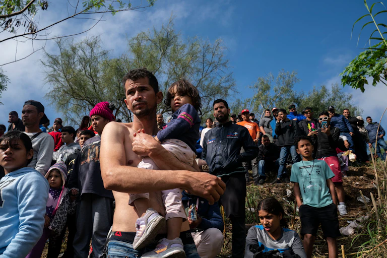 A barechested man holds his daughter as they join dozens of others waiting to receive asylum in the U.S.