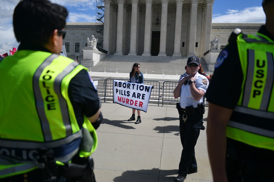A woman stands in front of a large white building with pillars and holds up a sign that say, 'abortion pills are murder.' Two people wearing yellow neon vests that say police stand nearby.