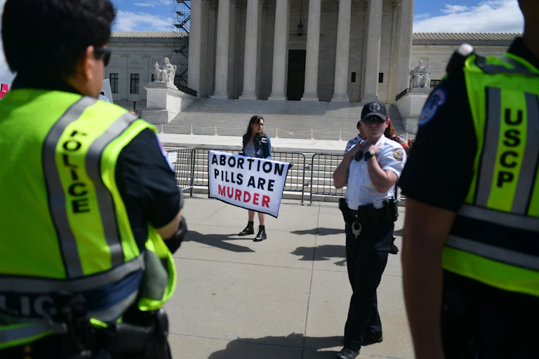 A woman stands in front of a large white building with pillars and holds up a sign that say, 'abortion pills are murder.' Two people wearing yellow neon vests that say police stand nearby.